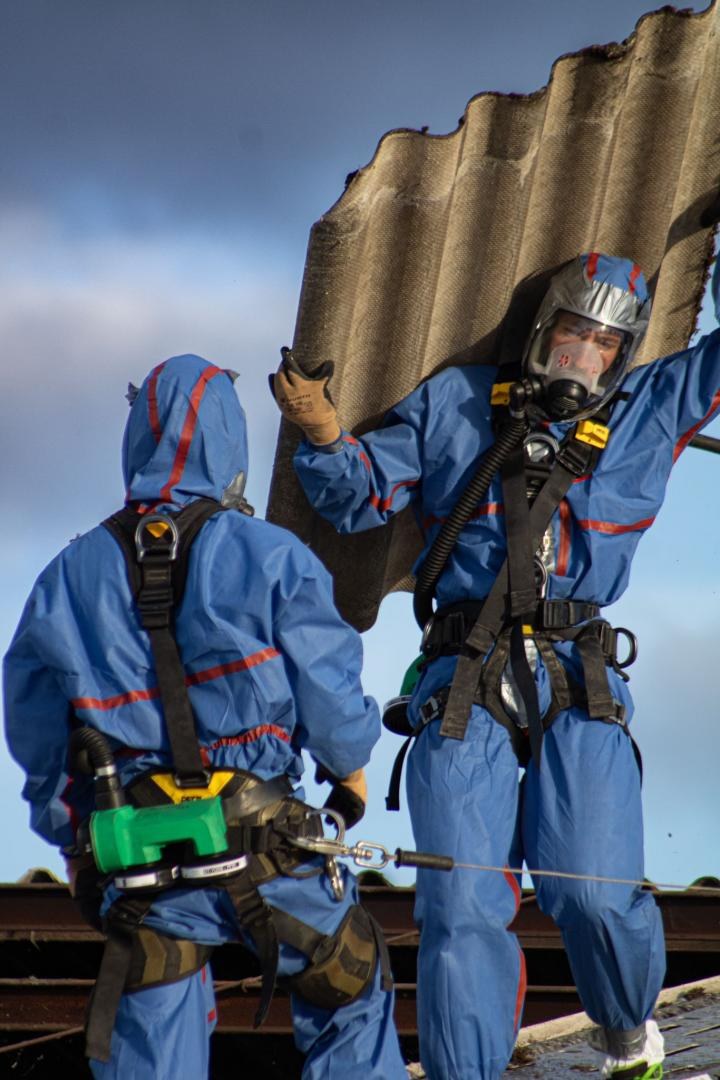 Two JP2R operators in protective suits and masks carrying a fibre-cement sheet removed from a roof